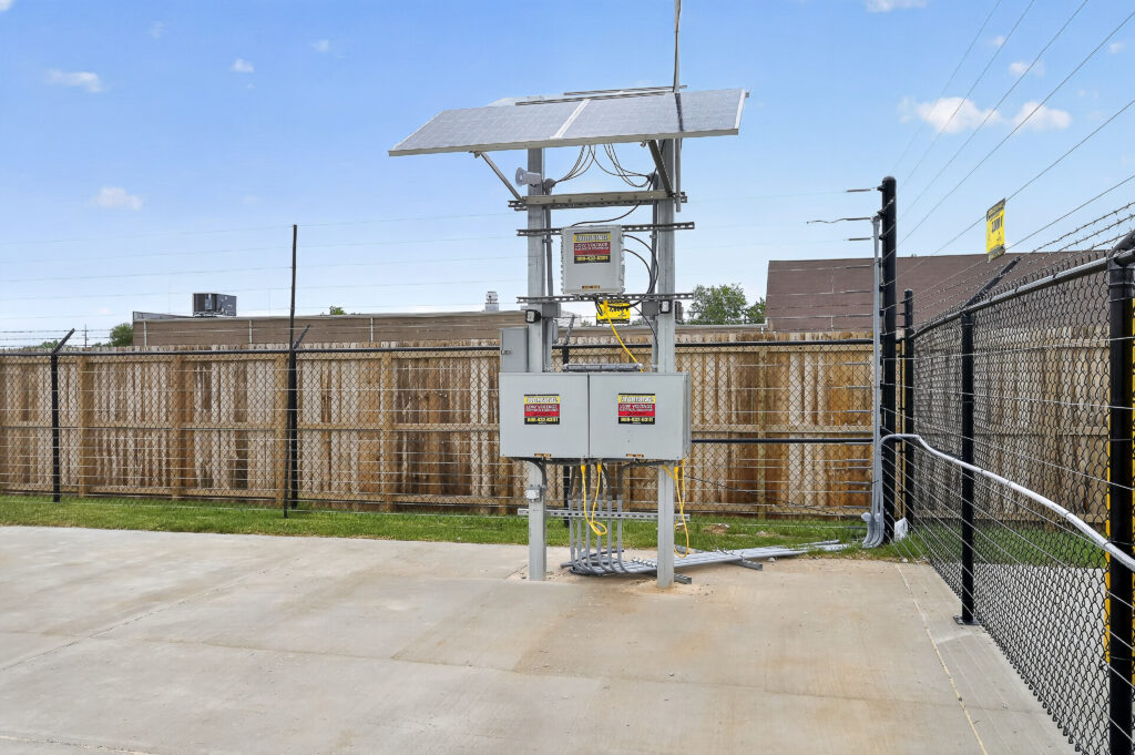 A solar-powered utility station with mounted electrical boxes and warning signs, situated on a concrete pad enclosed by a chain-link fence.