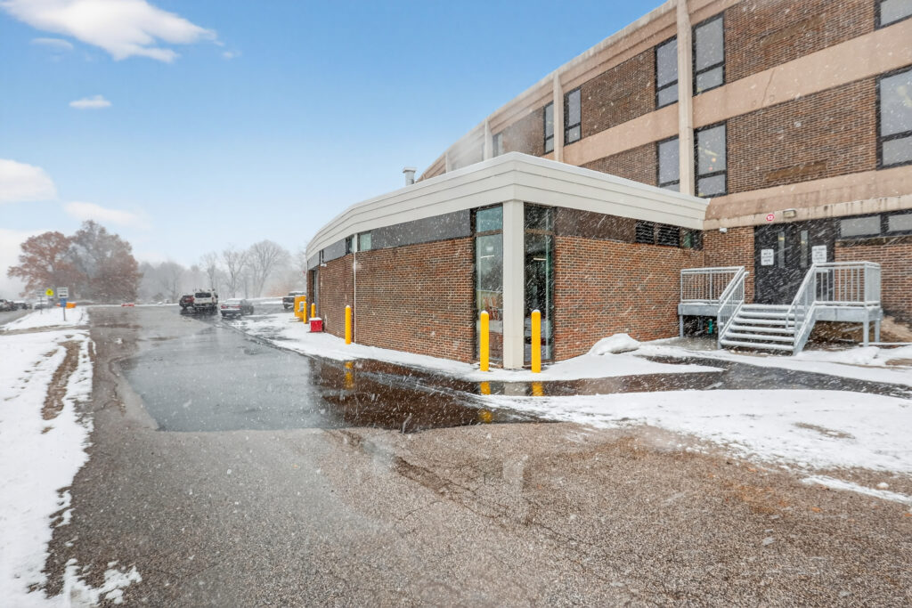 A brick building with a curved glass entrance during a light snowfall, featuring a set of stairs leading to a side door.