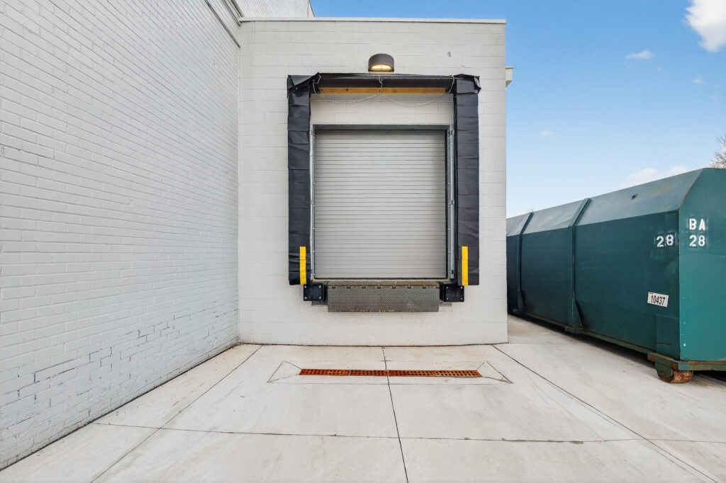 A loading dock door on a white brick building, next to a large green industrial dumpster.