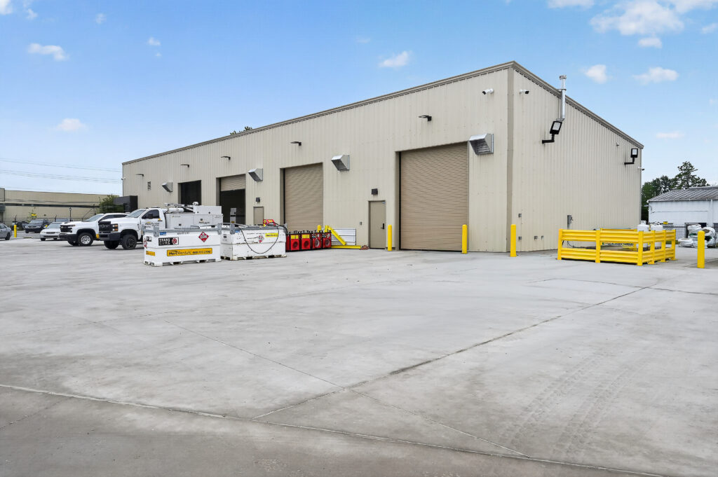 Tan industrial warehouse building with three large roll-up doors, parked service trucks, and equipment tanks in the paved front lot.