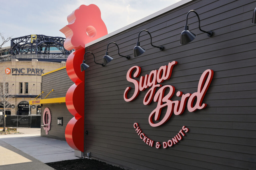 Exterior wall of Sugar Bird Chicken & Donuts restaurant, featuring a large red chicken-shaped accent and PNC Park in the background.