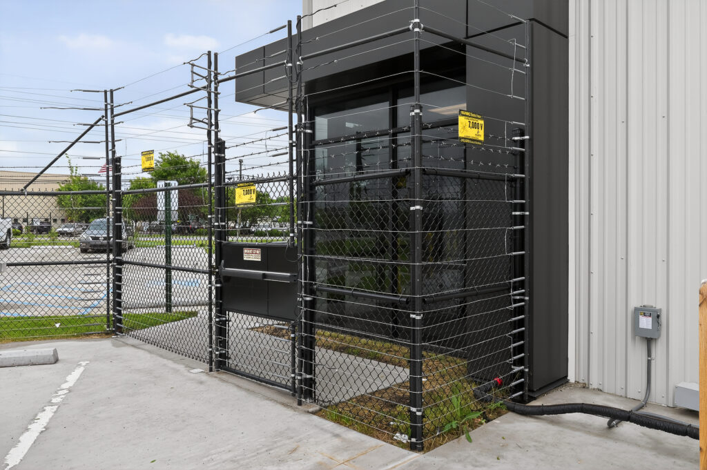 A glass building entrance surrounded by high chain-link fencing topped with barbed wire and electrical warning signs.