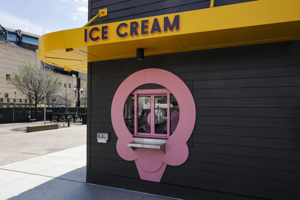 An ice cream stand with a yellow sign and a pink, ice-cream-shaped service window on a dark gray exterior wall.