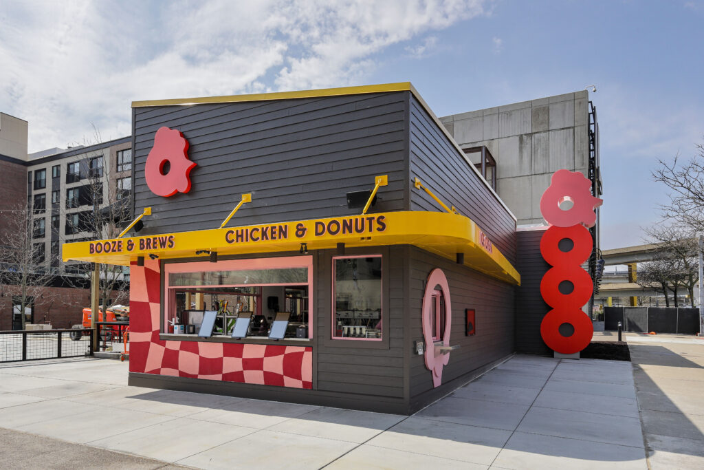 Exterior of a small takeout restaurant named “Chicken & Donuts” with a dark gray building, yellow awning, and red, pink, and white checkered accents.