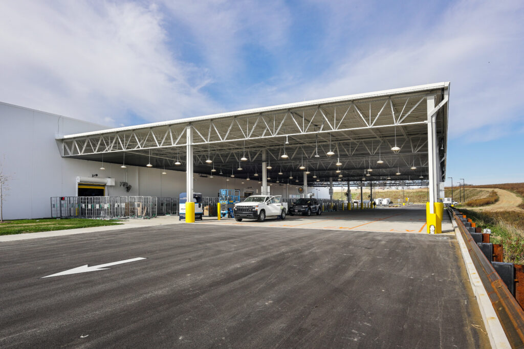A large warehouse loading dock area with a wide metal canopy, parked trucks, and wire storage cages.