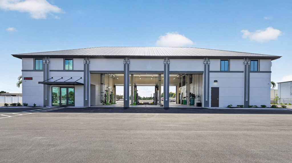 Exterior of a modern commercial automotive garage with multiple open bays and a large asphalt parking lot in the foreground.