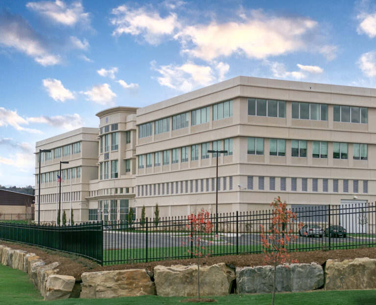 A multi-story beige office building with a black metal fence and stone wall in the foreground.