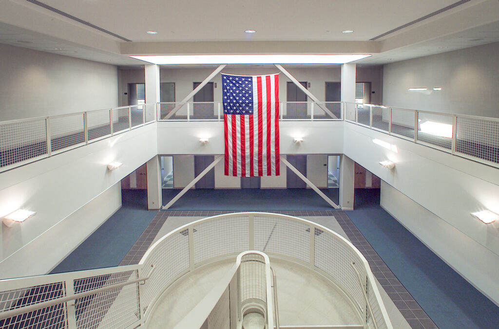 A large American flag hangs in the center of an open, multi-level building atrium with a central spiral staircase.