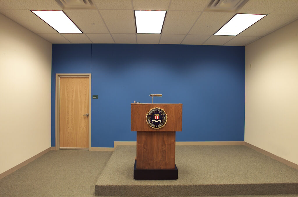 A wooden podium with the FBI seal on a carpeted platform in front of a blue wall and a closed door.