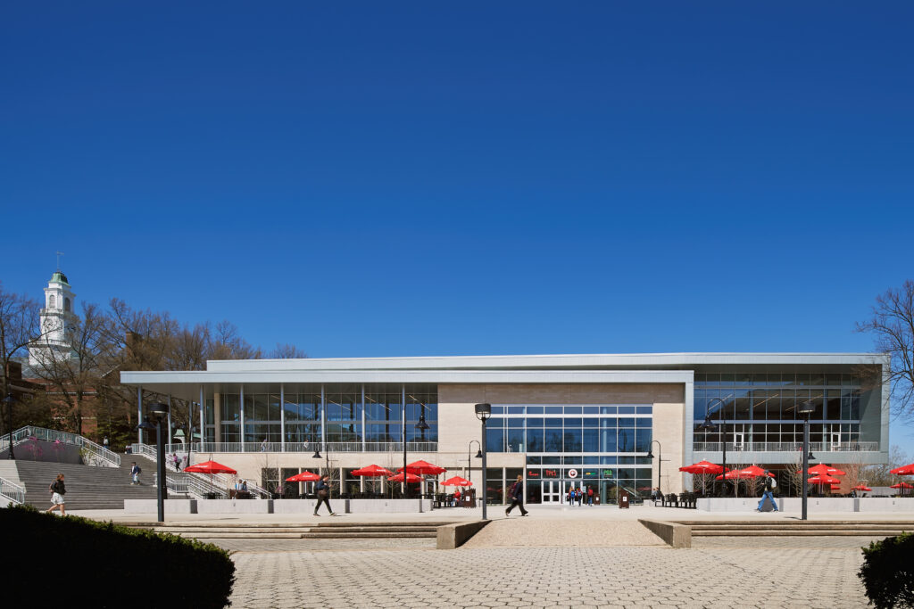 Modern building with large windows and a red-umbrella patio, located at a campus plaza.
