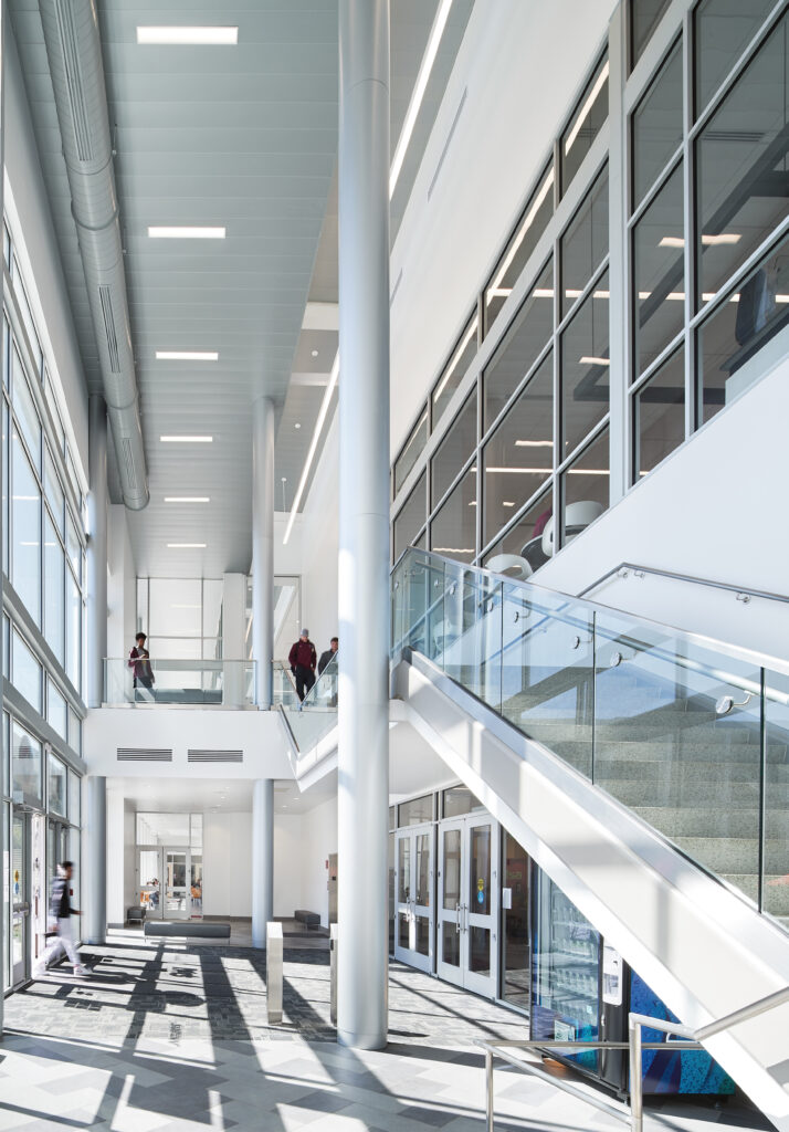 Modern building interior with high ceilings, glass-walled staircases, and people walking through the lobby.
