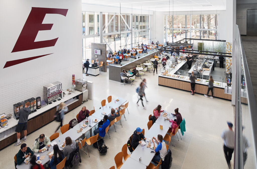 A high-angle view of a bright, modern university dining hall filled with students eating at tables and selecting food from a service counter.