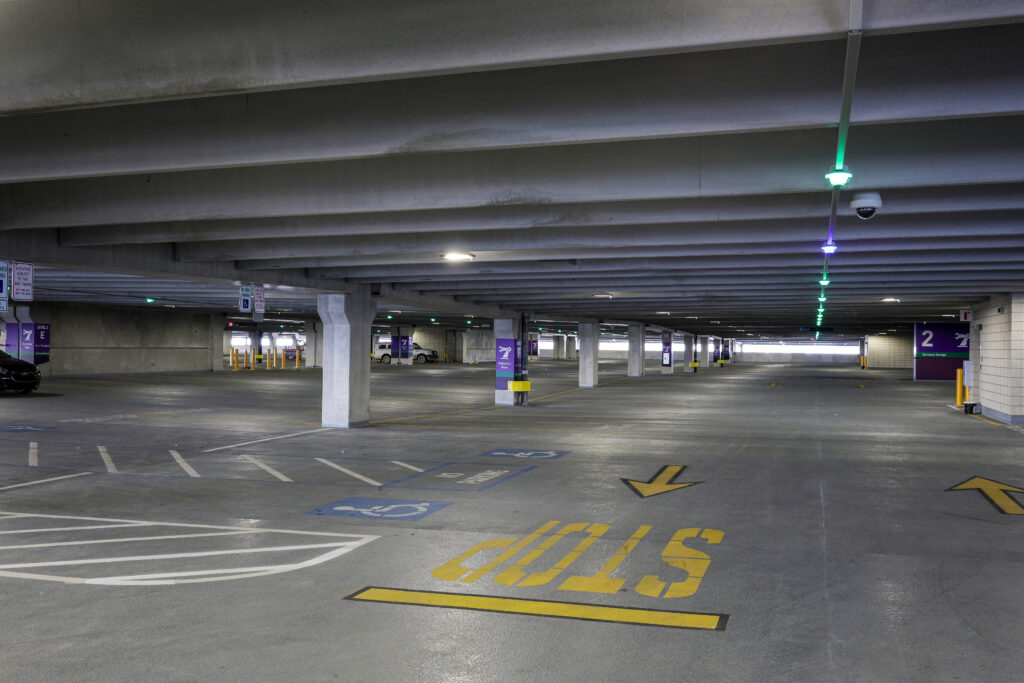 An empty parking garage with concrete pillars, marked parking spaces, and yellow directional arrows painted on the floor.