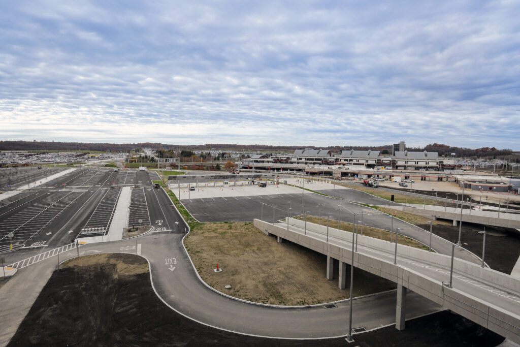 An aerial view of an airport terminal building with surrounding parking lots, access roads, and an elevated concrete ramp under a cloudy sky.