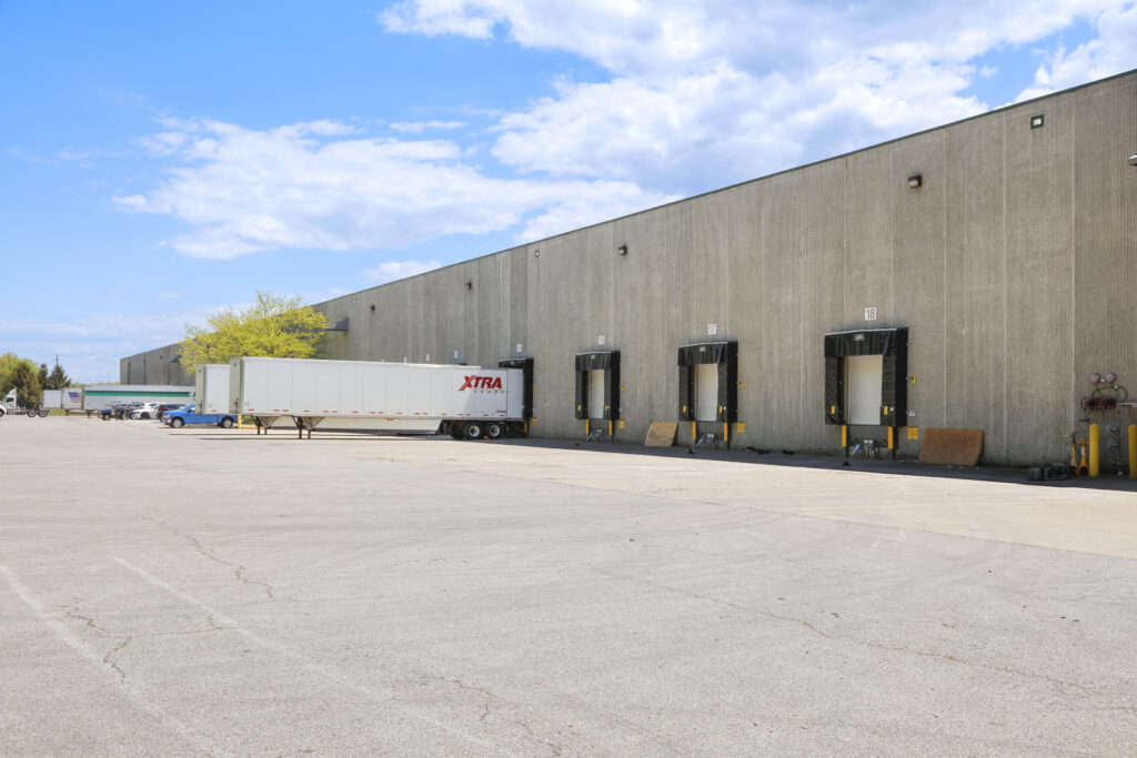 A semi-trailer truck parked at a loading dock outside a large warehouse building.