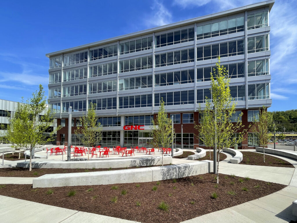 A multistory glass and brick office building with a GNC sign above the entrance and red outdoor furniture in the courtyard.