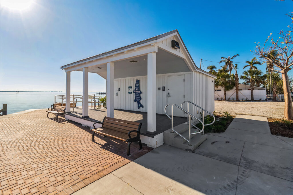A white public restroom building with a covered porch and benches on a brick waterfront plaza.