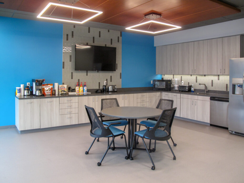 Office breakroom with a round table, four blue and black chairs, light-wood cabinets, a kitchen counter with appliances, and a wall-mounted television.