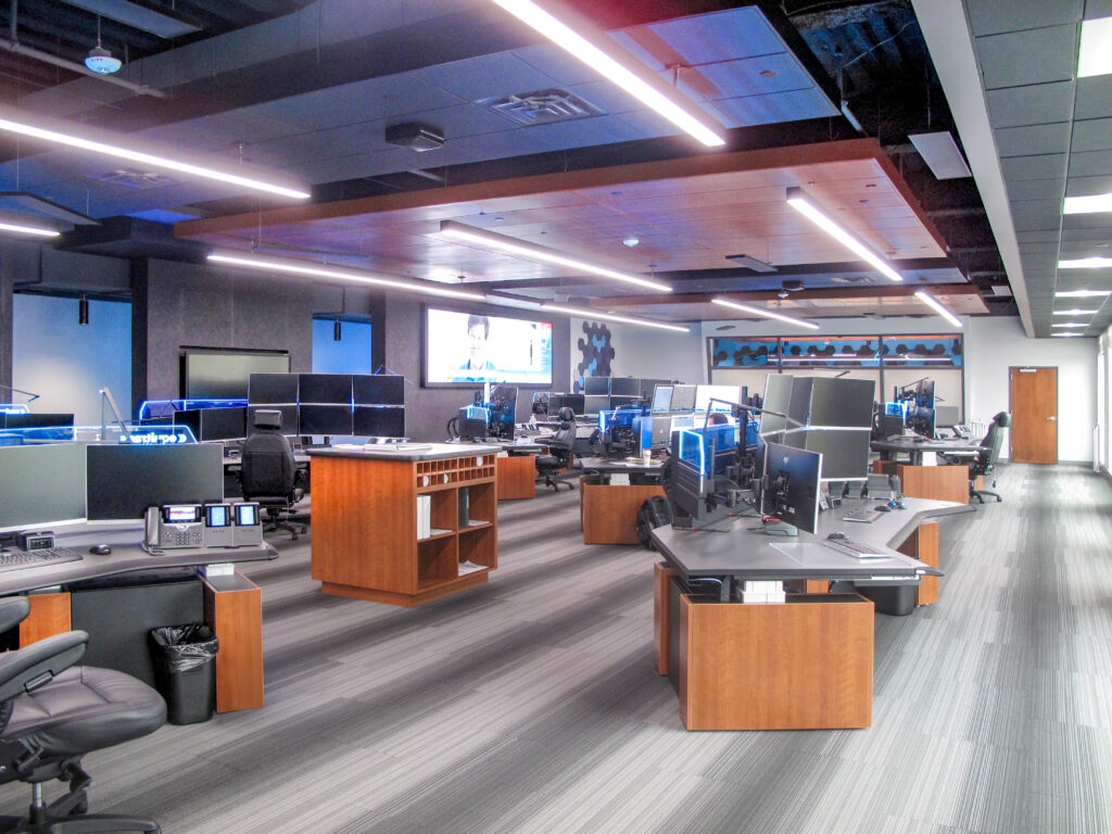 Modern office workspace with multiple rows of wooden desks equipped with computer monitors and office chairs.