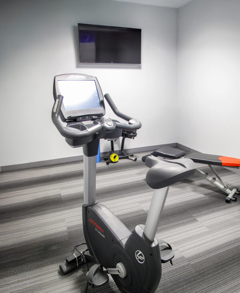 A stationary exercise bike in a small gym with a weight bench and a wall-mounted television.