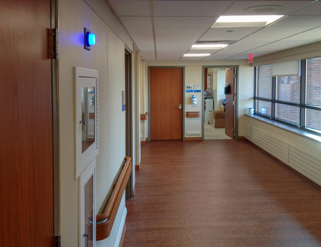 A hospital corridor with wood-look flooring, wall handrails, and several doorways, including an open door leading to a room with supplies.
