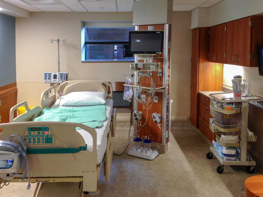 A hospital room with a patient bed, medical equipment mounted on a central pillar, and a supply cart next to wooden cabinets.