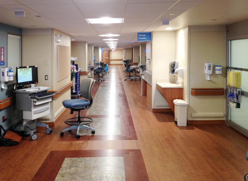 A hospital hallway featuring a mobile computer station, a desk chair, and a nurse station sign.