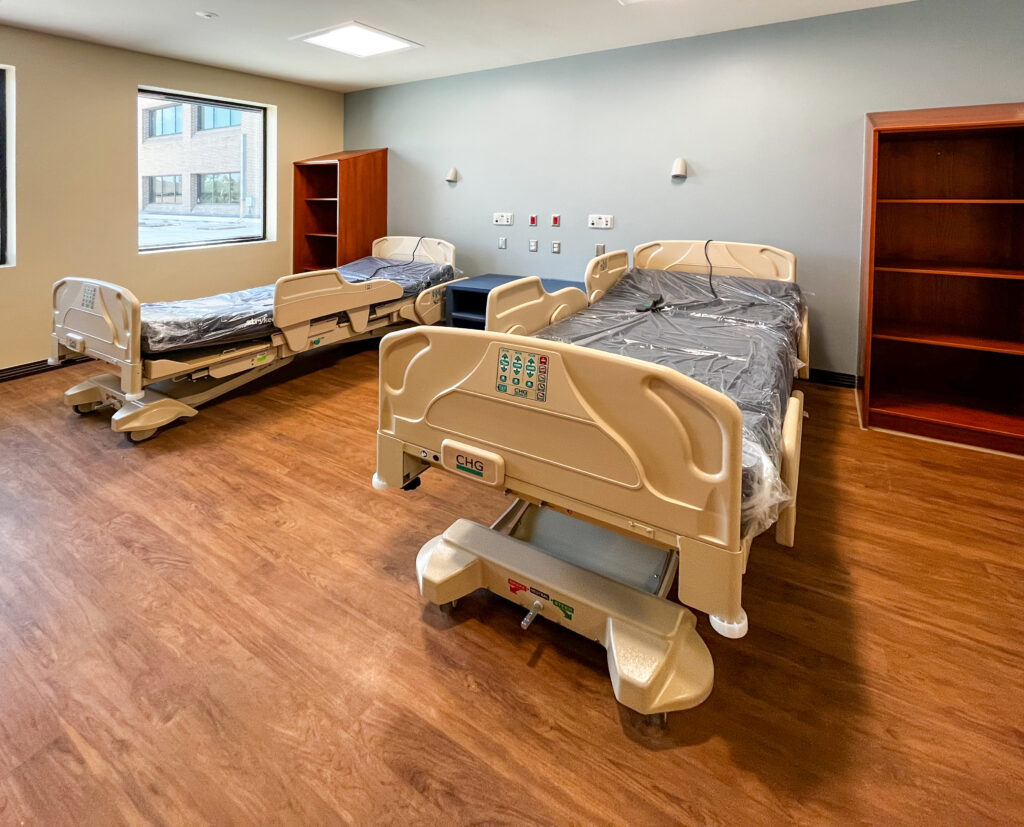 Two hospital beds covered in plastic in a room with wood-look flooring and two wooden bookshelves.