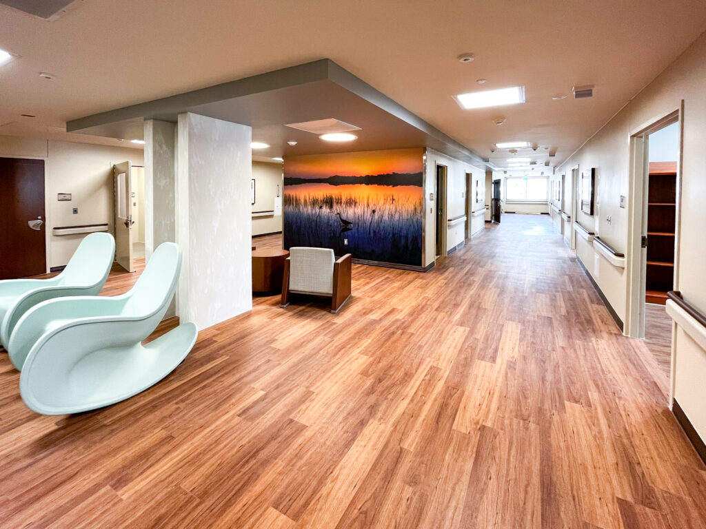 A hallway in a healthcare facility with wood-look flooring, two light-colored rocking chairs, an accent wall with a sunset marsh mural, and doorways leading to patient rooms.