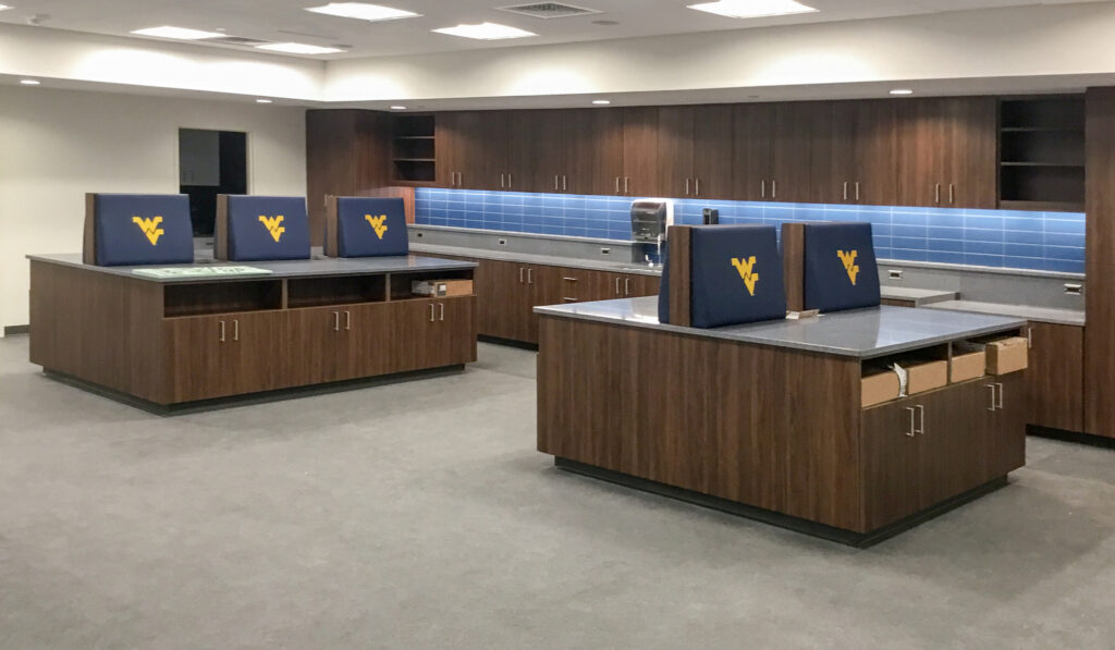 A modern breakroom with wood cabinets, gray countertops, and chairs featuring the West Virginia University logo.