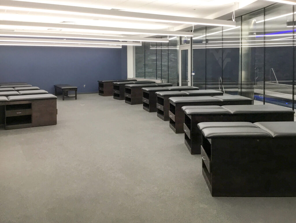 Rows of dark therapy tables with storage compartments in a clinical room with blue walls and floor-to-ceiling windows.