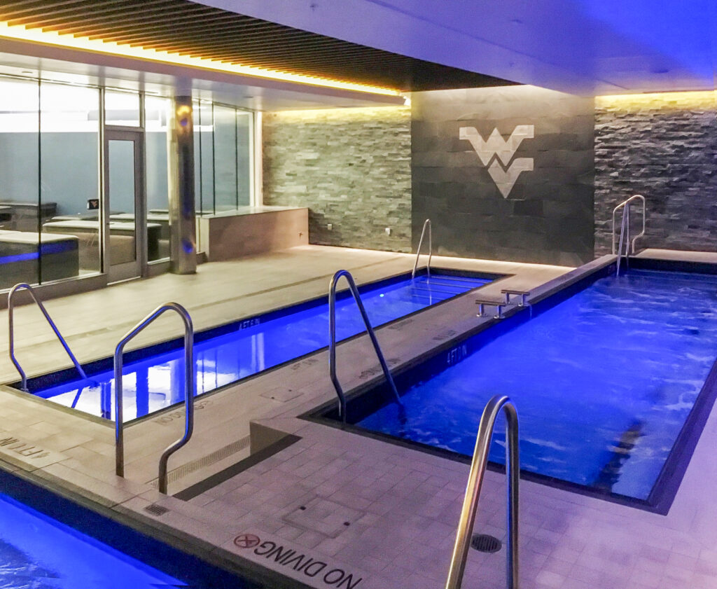 Two parallel indoor therapy pools with blue lighting and metal handrails, featuring a West Virginia University logo on the back stone wall.