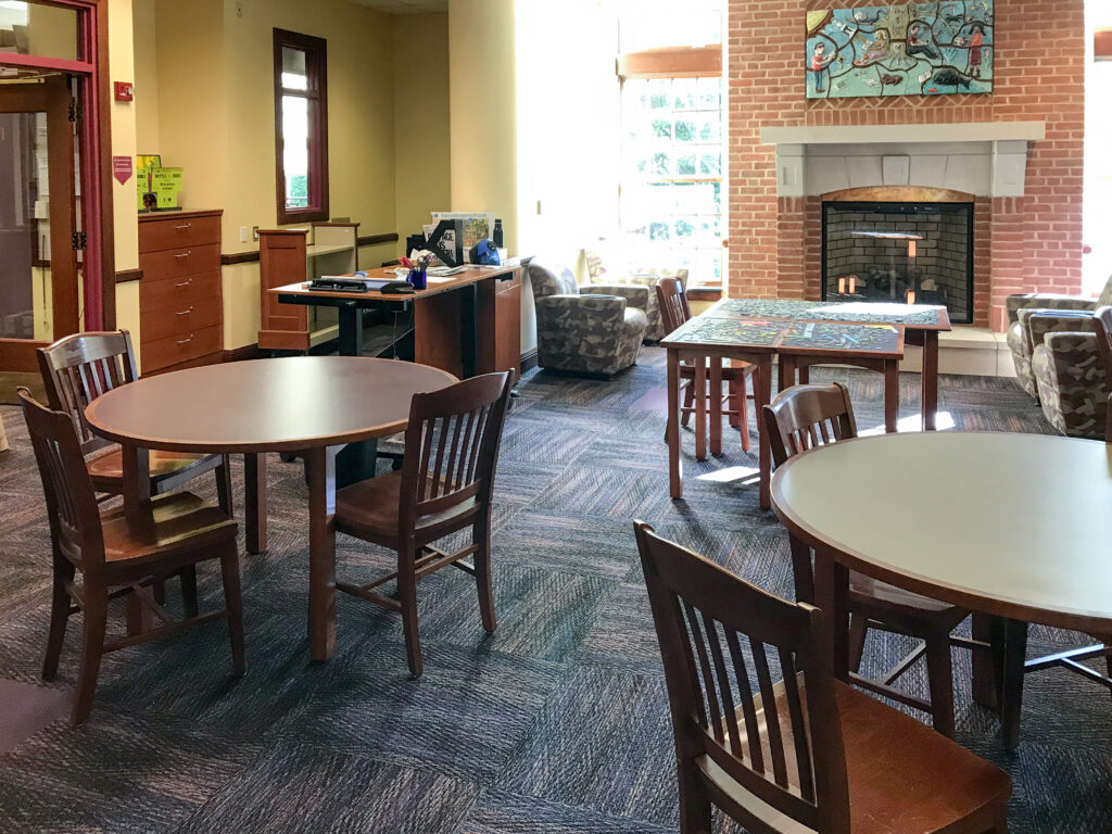 A library reading room with wooden tables, chairs, a fireplace with artwork above it, and a study desk.