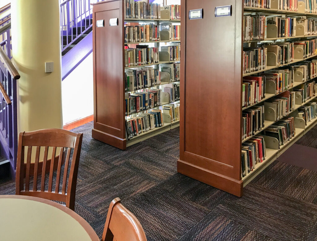 Two wooden bookshelves in a library, viewed from a seated area with chairs.