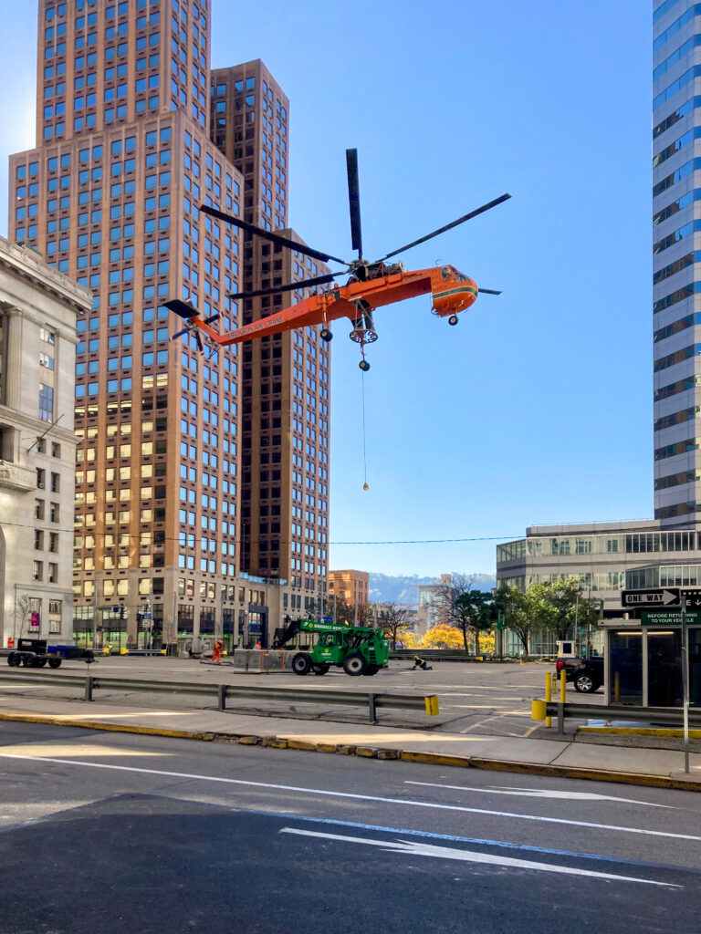 An orange Erickson Air-Crane helicopter hovers over a city plaza near a green construction forklift.