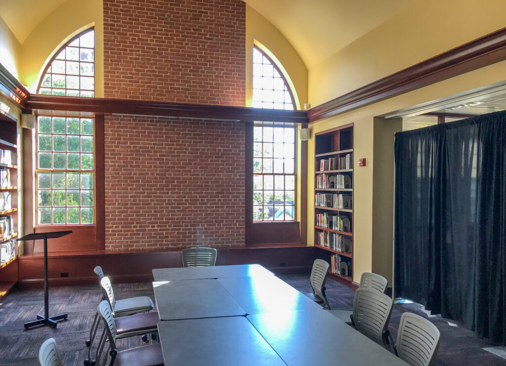 A library meeting room featuring a long conference table surrounded by chairs, with tall arched windows, a brick chimney breast, and built-in bookshelves.
