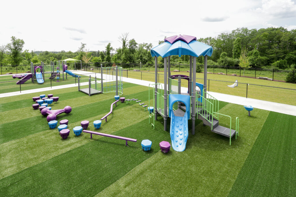 Outdoor playground with a blue and purple play structure featuring slides, climbing steps, and balancing pods on artificial turf.