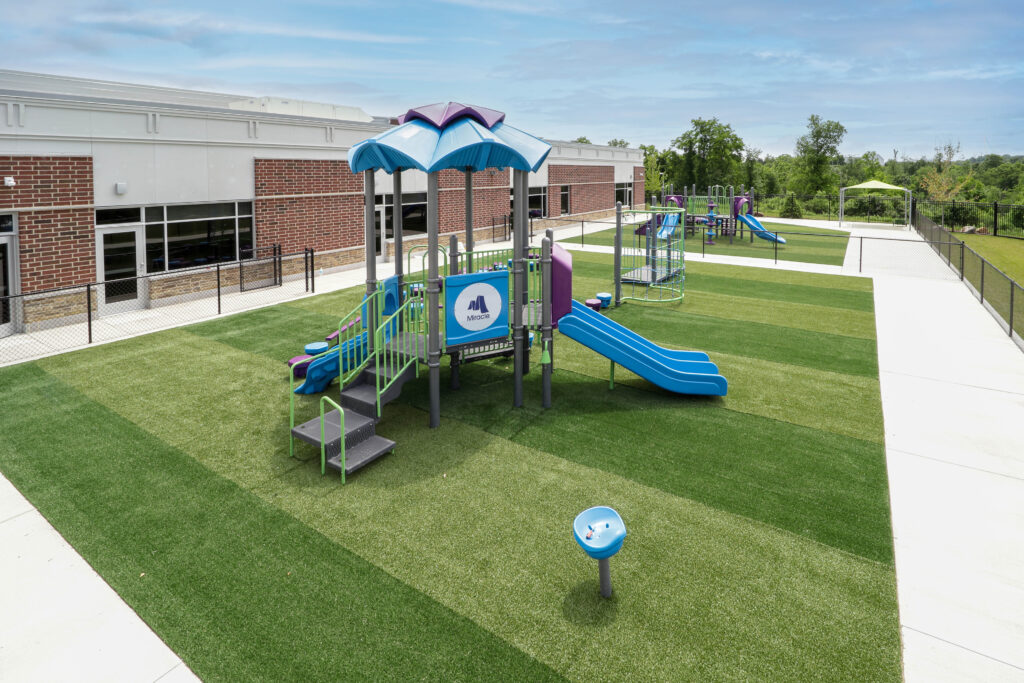 A playground with a Miracle brand structure featuring blue slides, set on a green turf field next to a brick building.