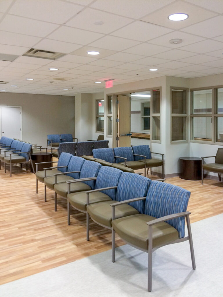 Empty rows of waiting room chairs arranged in an office space with wood-look flooring and interior windows.