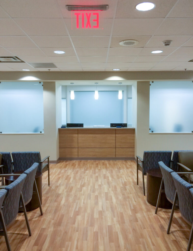 Reception desk in a waiting area with chairs, frosted glass panels, and an illuminated exit sign overhead.