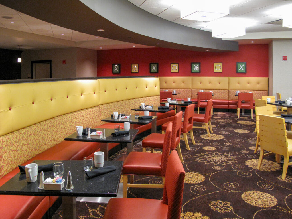 Dining area with long yellow booths, red chairs, and black tables set for service, in front of a red wall decorated with framed tic-tac-toe boards.