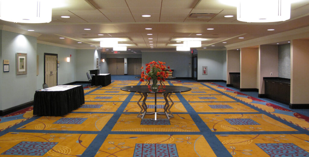 A long hotel conference hallway featuring a patterned yellow and blue carpet, a central round table with a vase of red flowers, and side tables covered with black cloths.