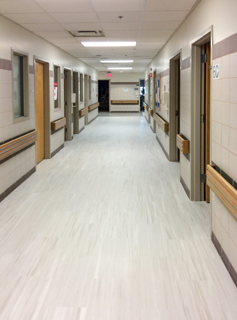 A long hospital hallway with tiled walls, wood-trimmed doors, and light-colored flooring.