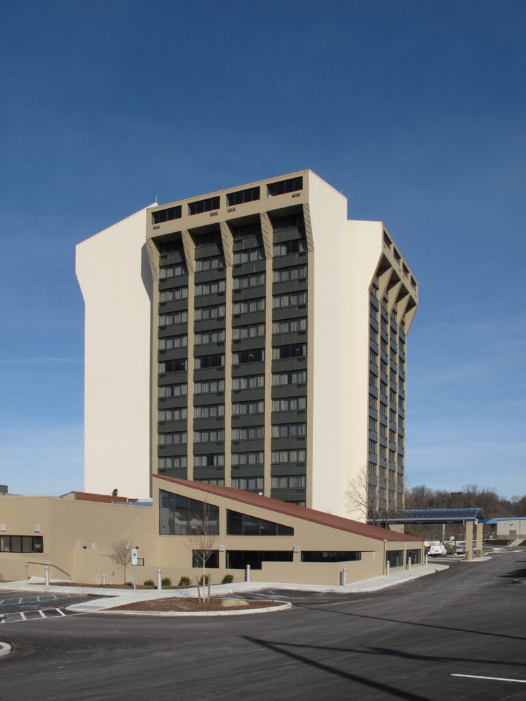 A high-rise hotel with a beige facade and recessed dark windows, featuring a lower, angled building wing extending into the parking lot.