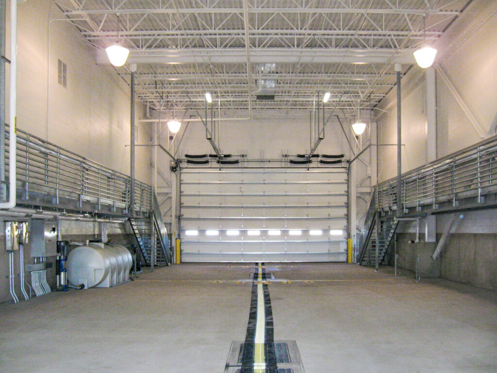 An empty industrial warehouse bay with a large overhead door, elevated side walkways, and a central floor drain.