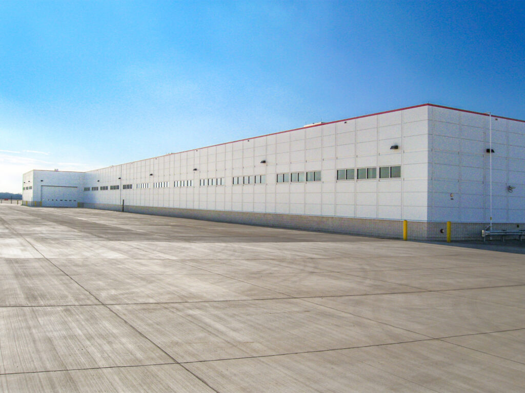 A long, modern white industrial warehouse building with a large concrete lot in the foreground under a clear blue sky.