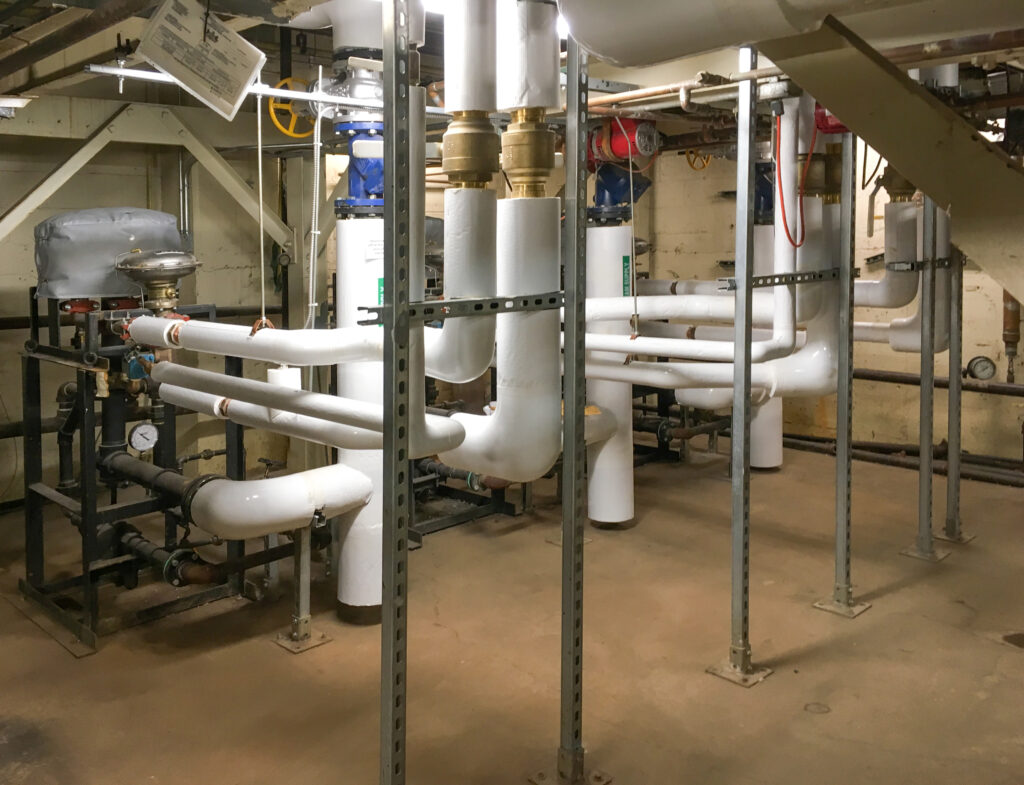 Industrial mechanical room with insulated white piping, vertical steel support beams, and various valves.