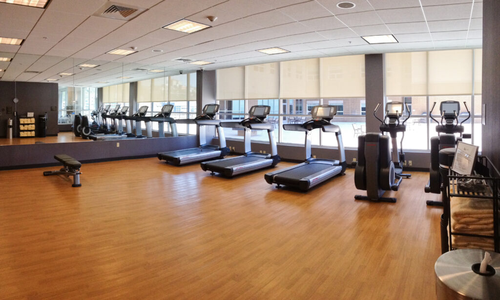 A row of treadmills and elliptical machines in a gym with wood floors and a mirrored wall.