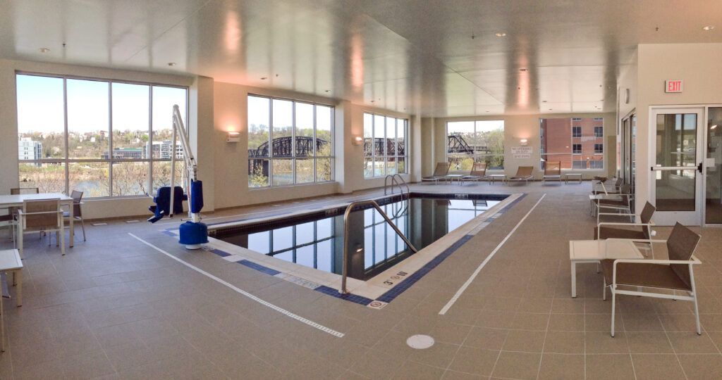Indoor swimming pool with a lift, lounge chairs, and large windows overlooking a bridge.