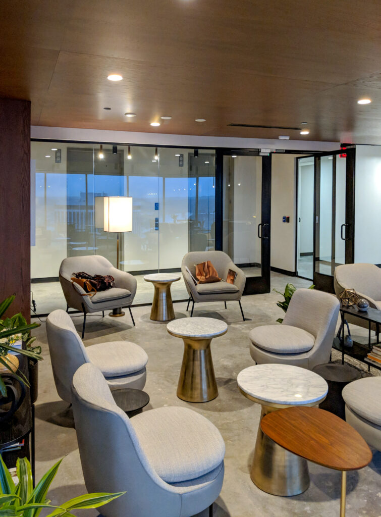 A lounge area with several gray armchairs, round side tables, and glass walls looking out to a cityscape.